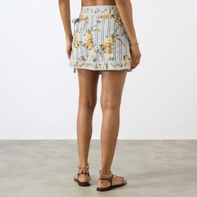 Model wearing Agua Bendita Luna Valle Mini Skirt in multicolour floral stripe print with tie waist and scalloped hem, styled with tan strappy sandals.