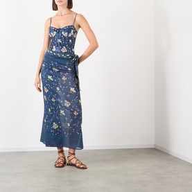 Model wearing Agua Bendita multicolour floral swimsuit styled as dress with wrap skirt overlay, paired with brown lace-up flat sandals.