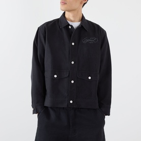 Model wearing black cropped utility jacket with embroidered chest detail, white buttons, and front patch pockets, styled with matching black trousers