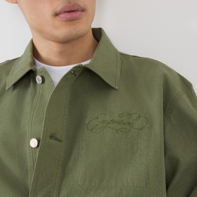 Model wearing Emporium 89 Ripstop Logo Jacket in green, showing tonal logo embroidery on chest, point collar, and white tee underneath.