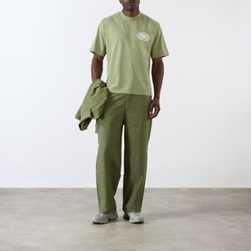 Model wearing Emporium EE Logo T-Shirt in khaki with matching wide-leg trousers and grey trainers, carrying an overshirt.