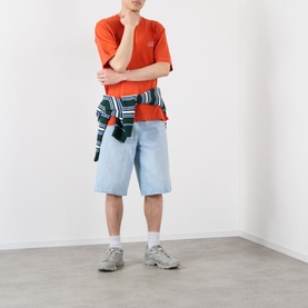 Model wearing Emporium Script Logo T-Shirt in orange, styled with light wash denim shorts, striped shirt tied at waist, and grey trainers.