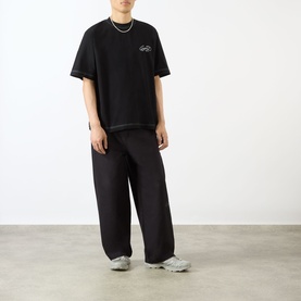 Model wearing Emporium Script Logo T-Shirt in black with embroidered logo, styled with wide-leg black trousers and grey trainers.