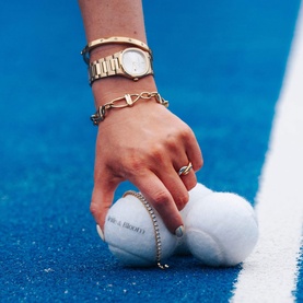 Model with Mettle & Bloom gold bracelet, styled with a watch and ring, hand reaching for tennis ball on blue court.