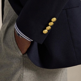 Close-up of navy blazer sleeve with gold embossed buttons, styled with grey trousers and striped shirt cuff visible underneath