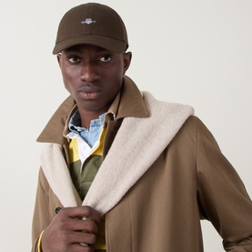 Model with Gant Shield Logo Baseball Cap in Shadow Brown, six-panel cotton cap with embroidered logo, styled with tan coat and layered shirts