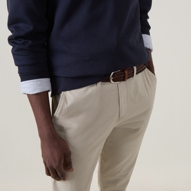Model wearing Gant Clay Beige cotton chino trousers, styled with a navy sweatshirt, striped shirt, and tan leather belt with gold buckle.
