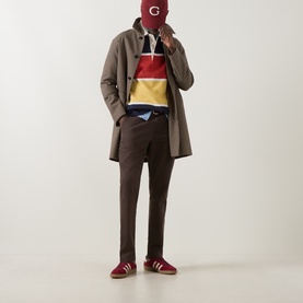 Model wearing Gant Shadow Brown chino trousers, styled with a striped rugby polo, grey overcoat, and red suede trainers.