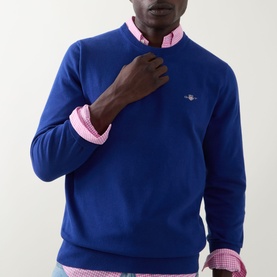 Model wearing Gant Shield Logo Sweater in Crisp Blue crew neck cotton knit, styled with a pink checked shirt underneath.