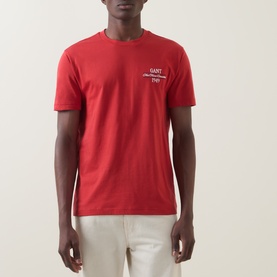 Model wearing Gant 1949 Logo T-Shirt in Maple Fire red, with small chest logo print, crew neck and short sleeves, styled with cream trousers.