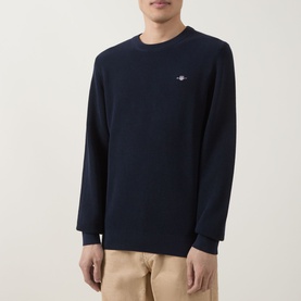 Model wearing Gant Shield Logo Micro Textured Sweater in Evening Blue, crew neck with embroidered shield logo, styled with beige trousers.