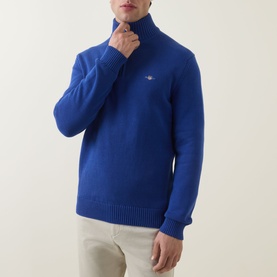 Model wearing Gant Shield Logo Half-Zip Sweater in crisp blue with embroidered shield logo, half-zip funnel neck, styled with cream trousers.