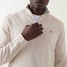 Model wearing Gant Shield Half-Zip Sweater in clay beige, showing embroidered shield logo and half-zip neckline detail, styled over white crew neck.