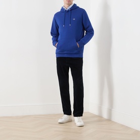 Model wearing Gant Shield Drawstring Hoodie in Crisp Blue with kangaroo pocket, styled with black trousers and white trainers.