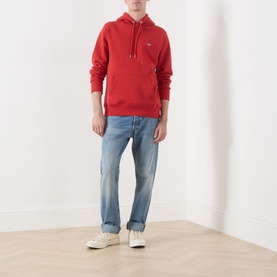 Model wearing Gant Shield Drawstring Hoodie in Maple Fire with kangaroo pocket and drawstring hood, styled with light wash jeans and white trainers.