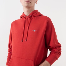 Model wearing Gant Shield Drawstring Hoodie in Maple Fire red, showing embroidered shield logo, drawstring hood, and kangaroo pocket.