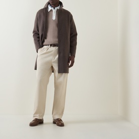 Model wearing Gant Rugby Knitted Polo Shirt in Mist Taupe, styled with dark brown overcoat, cream trousers and brown loafers.