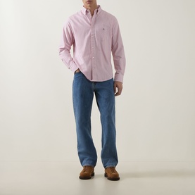 Model wearing Gant Shield Logo Striped Shirt in Maple Fire, pink stripes, button-down collar, styled with blue jeans and tan shoes.