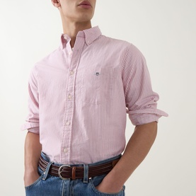 Model wearing Gant Shield Logo Striped Shirt in Maple Fire, pink stripe, button-down collar, long sleeves, styled with blue jeans and brown belt.