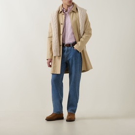 Model wearing Gant Shield Logo Striped Shirt in Maple Fire, styled with a beige mac coat, blue jeans, and brown suede shoes.