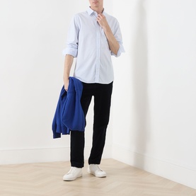 Model wearing Gant Classic Stripe Banker Shirt in light blue with black trousers, white trainers, and a blue jacket draped over one arm.