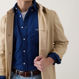 Model wearing Gant Dark Indigo Reg Shirt in 100% cotton, styled under a beige mac coat with jeans and brown belt.