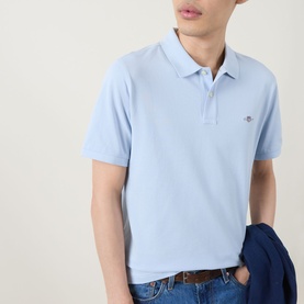 Model wearing Gant Regular Polo in Fresh Blue, short sleeve pique cotton with logo badge, styled with blue jacket and brown belt jeans.