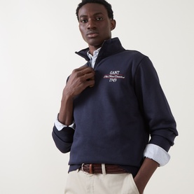 Model wearing Gant Evening Blue half-zip sweatshirt with embroidered logo, styled with a white shirt and beige chinos.