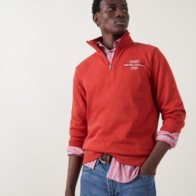 Model wearing Gant Maple Fire half-zip sweatshirt with logo chest embroidery, styled over striped shirt with jeans.
