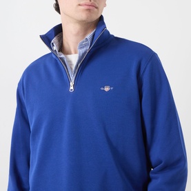 Model wearing Gant crisp blue half-zip sweatshirt with embroidered logo, styled over a striped shirt collar visible at neckline.