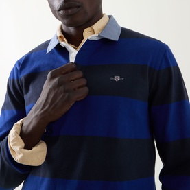 Model wearing Gant Shield Logo Striped Rugby Shirt in crisp blue, navy block stripes, contrast chambray collar, embroidered chest logo visible.