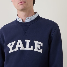 Model wearing Gant V-Stitch Logo Sweatshirt in navy with Yale graphic, styled with a blue striped shirt collar visible at neckline.