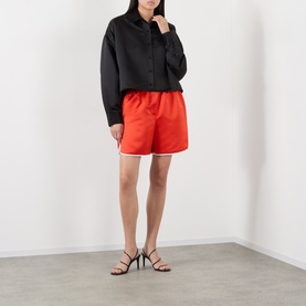 Model wearing The Frankie Shop black cropped polyester shirt, styled with red shorts and black heeled sandals.