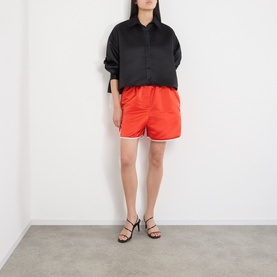 Model wearing The Frankie Shop black oversized polyester shirt, styled with red satin shorts and black strappy heeled sandals.