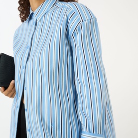 Model wearing The Frankie Shop Oana Padded Stripe Shirt in Blue Multi Stripe, with padded dropped shoulders, styled with a black clutch bag.