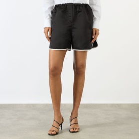 Model wearing The Frankie Shop Adia Piped Shorts in black satin with white contrast piping and elasticated waistband, styled with strappy heeled