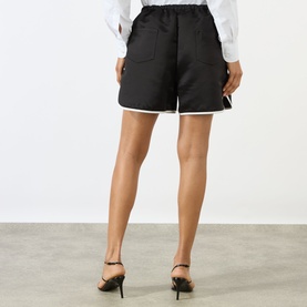 Model wearing The Frankie Shop Adia Piped Shorts in black satin with white contrast piping, elasticated waist and patch pockets, styled with black