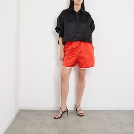 Model wearing The Frankie Shop red satin shorts with white trim, styled with black satin shirt and black strappy heels.
