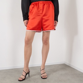 Model wearing The Frankie Shop red satin shorts with white trim, styled with a black long-sleeve top and black heeled sandals.