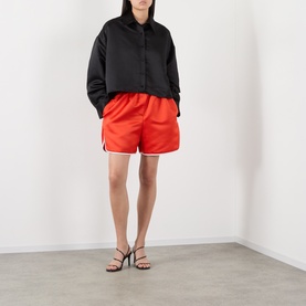 Model wearing The Frankie Shop red satin shorts, styled with a black oversized shirt and black strappy heeled sandals.