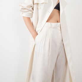 Model wearing The Frankie Shop ivory silk wide-leg trousers with deep pleats, styled with a matching ivory trench coat and black bra top.