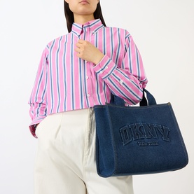 Model carrying DKNY Hadlee Medium Tote Bag in dark denim with embossed logo, top handles, gold-tone hardware, styled with pink striped shirt