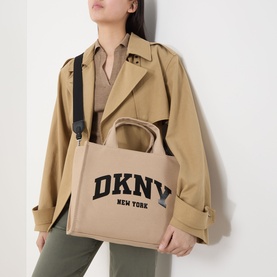 Model carrying DKNY Hadlee Medium Tote Bag in khaki and black canvas with logo branding and black adjustable shoulder strap, styled with trench coat
