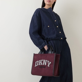 Model carrying DKNY Hadlee Medium Tote Bag in aged wine faux suede with logo branding and silver-tone hardware, styled with navy jumpsuit