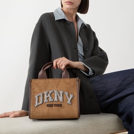 Model carrying tan structured tote bag with rhinestone DKNY logo lettering and short brown handles, styled with grey blazer and striped shirt