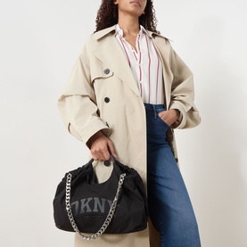 Model carrying DKNY Mabel Large Tote Bag in black with silver chain strap and logo detail, styled with beige trench coat and jeans.