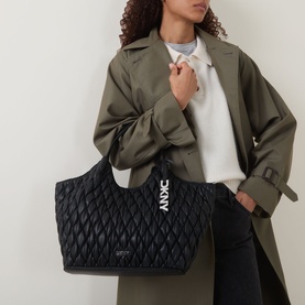 Model carrying DKNY Paula Quilted Medium Tote Bag in black with silver-tone hardware and logo branding, styled with an olive trench coat