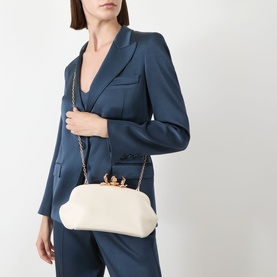 Model carrying small cream clutch bag with gold chain strap and embellished clasp, styled with blue tailored suit