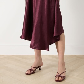 Model wearing Marella Libero Draped Dress in Bordeaux, satin midi length with asymmetric hem, styled with burgundy strappy heeled mules.