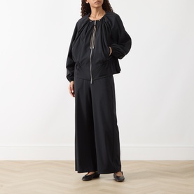 Model wearing Max&Co Moli Technical Blouse in black with two-way zip, drawstring neck, styled with wide-leg black trousers and flat shoes.